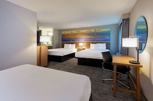 Quarto de hotel com duas camas, uma escrivaninha, uma cadeira, uma luminária de mesa, um espelho e um quadro de pôr do sol.