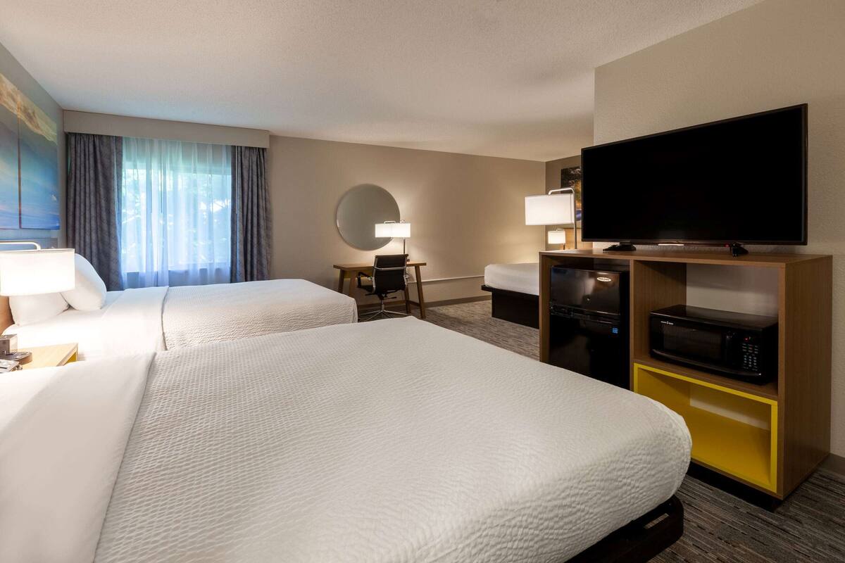 Une chambre d’hôtel avec deux lits, un bureau avec une chaise, un téléviseur à écran plat, un four à micro-ondes et une grande fenêtre avec des rideaux.