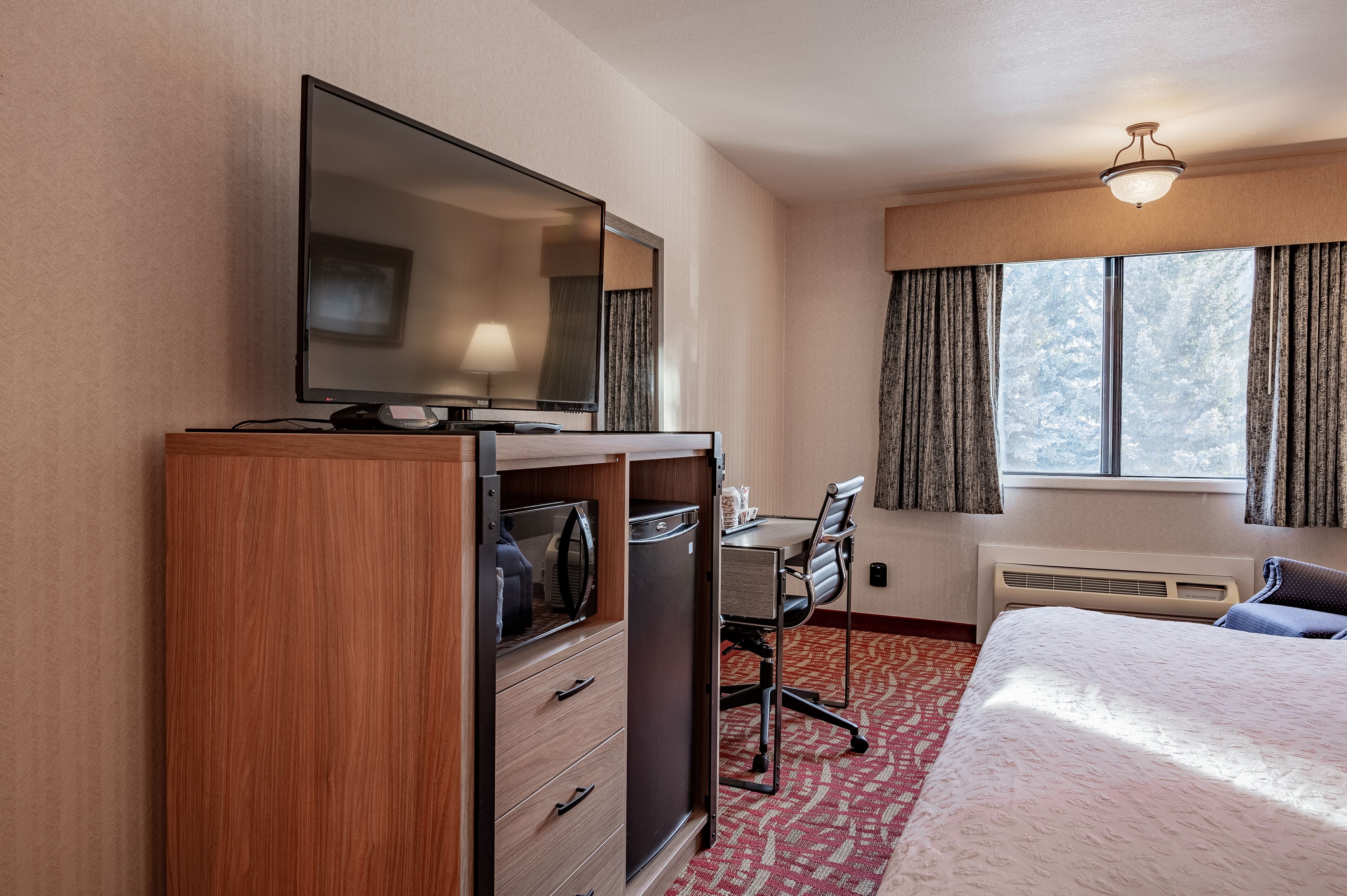 A hotel room with a bed, a desk with a chair, a wardrobe, a television, and a window with curtains.