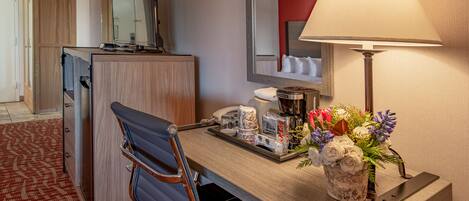 A hotel room with a wooden desk, a chair, a lamp, a telephone, a coffee maker, and a floral arrangement.