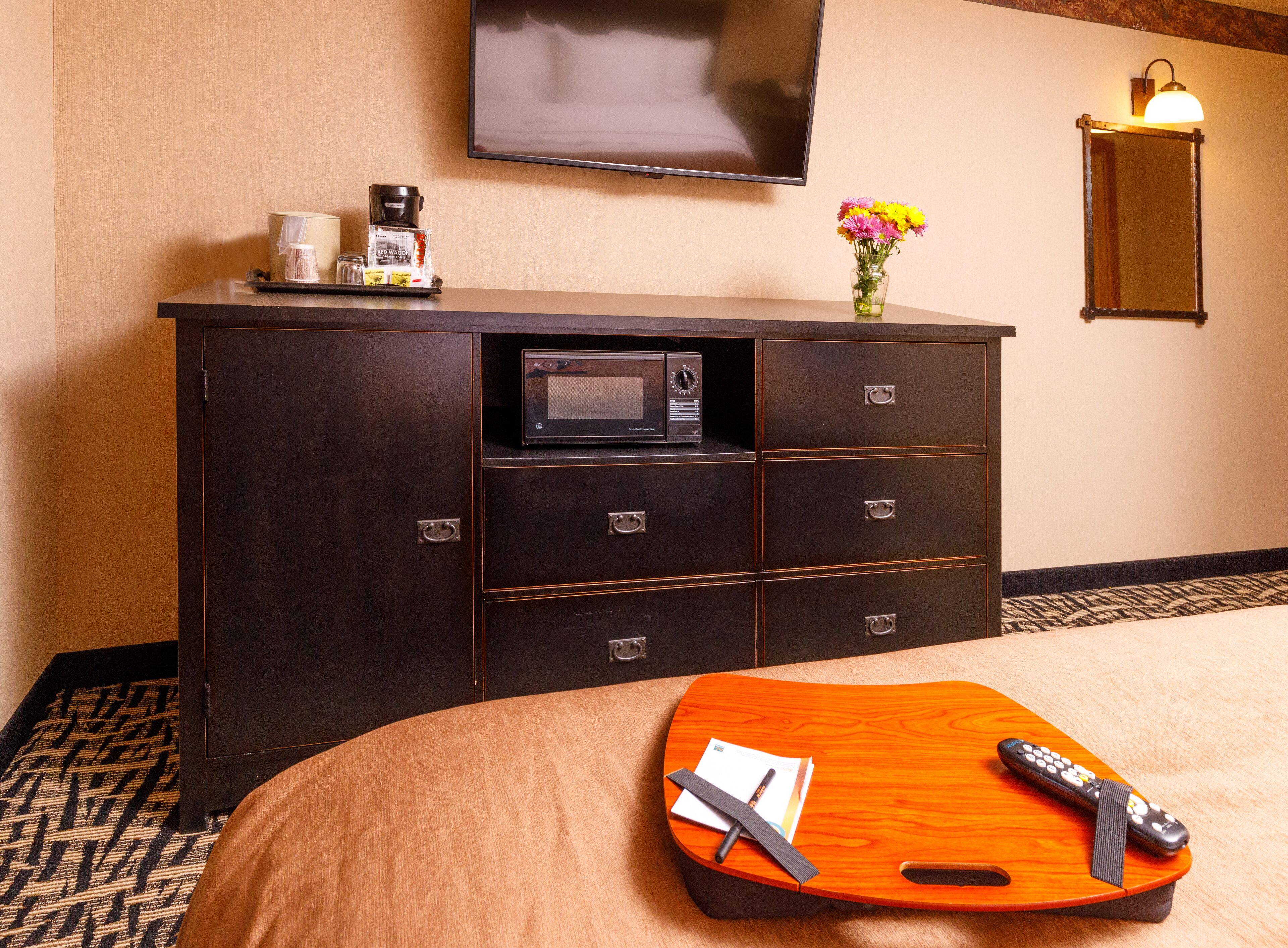 A hotel room with a dark wooden dresser, a flat-screen TV, a vase with flowers, and a wooden tray with a remote control and pen.