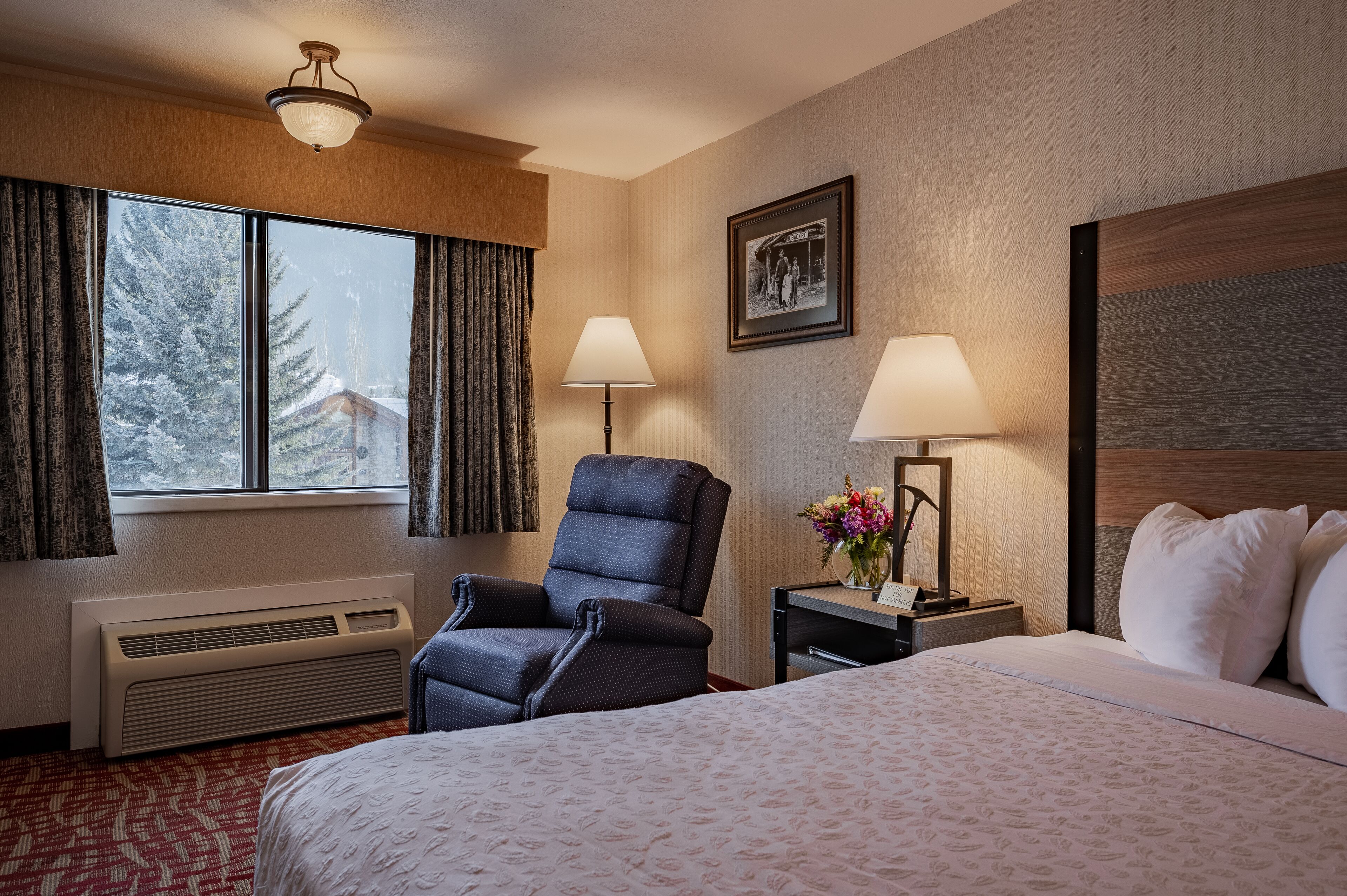 A hotel room with a bed, a chair, a desk with a lamp, a picture on the wall, and a window with curtains.
