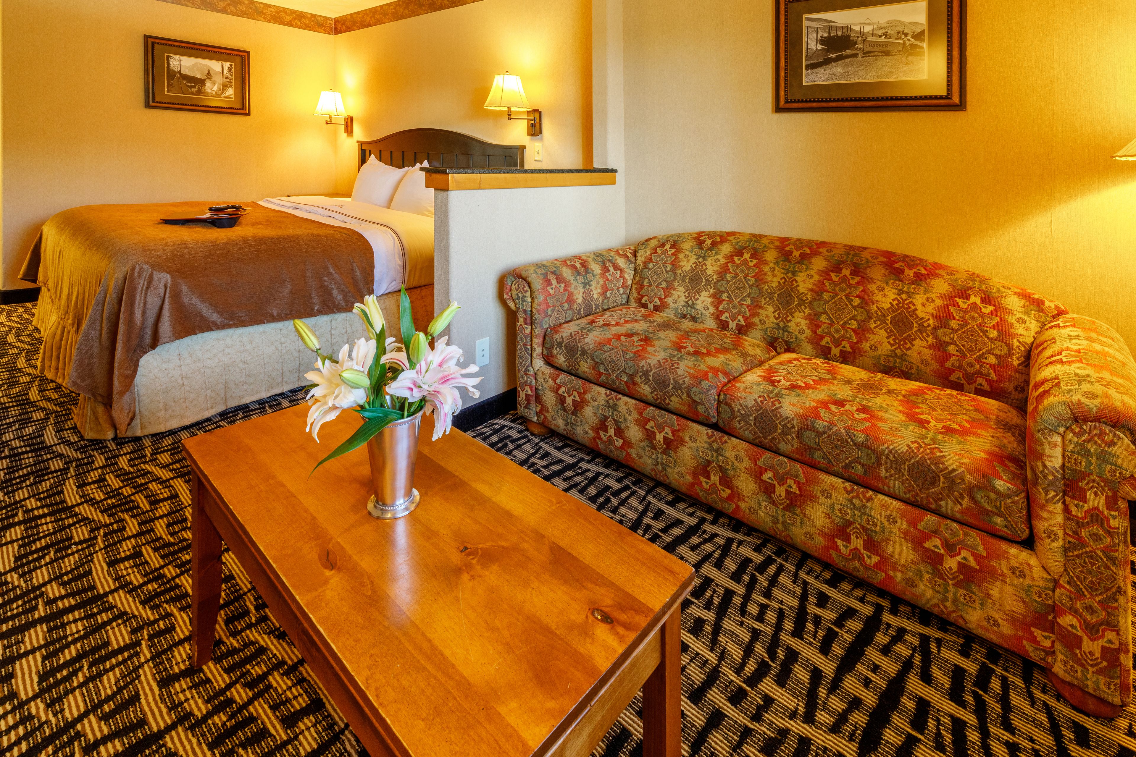 A hotel room with a bed, a sofa, a wooden table with a vase of flowers, and patterned carpet.