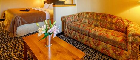 A hotel room with a bed, a sofa, a wooden table with a vase of flowers, and patterned carpet.