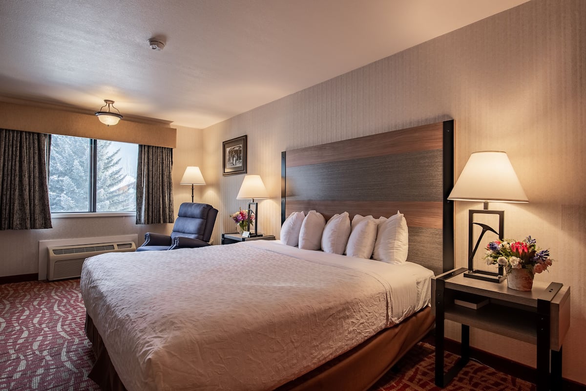 A hotel room with a large bed, two bedside lamps, a chair, a window with curtains, and a framed picture on the wall.