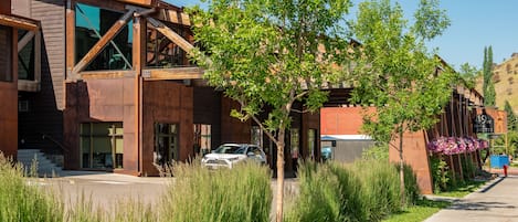 Un edificio moderno con una amplia entrada de vidrio, un árbol en primer plano y un coche estacionado afuera.