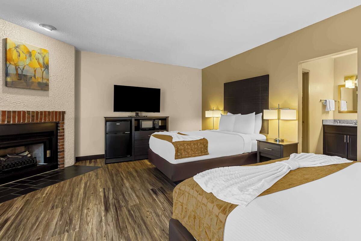 A hotel room with a fireplace, a flat-screen TV, two beds with white linens, a nightstand with a lamp, and a bathroom with a mirror and towel rack.