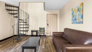 Ein Wohnzimmer mit einer braunen Ledersofa, Holzboden, einem schwarzen Tisch, einem gemusterten Sessel, einer Wendeltreppe und einer Tür mit einem Gemälde an der Wand.