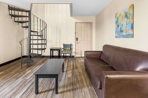 A living room with a brown leather sofa, a wooden floor, a black table, a patterned chair, a spiral staircase, and a door with a painting on the wall.