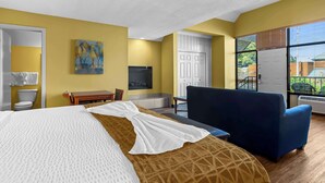 Ein Hotelzimmer mit einem Bett, einem blauen Sessel, einem Schreibtisch, einem Gemälde, einer weißen Tür und großen Fenstern.