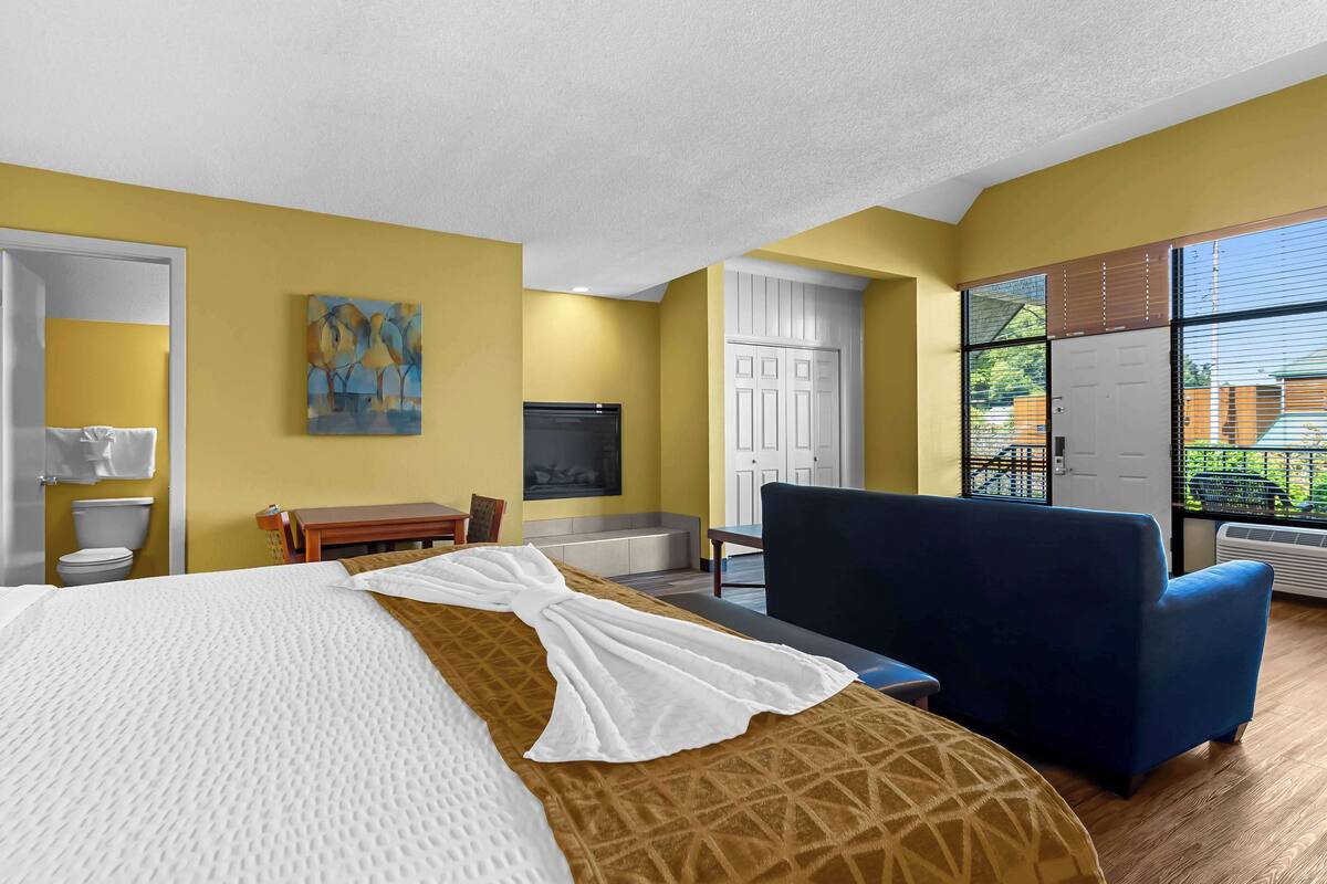 A hotel room with a bed, a blue armchair, a desk, a painting, a white door, and large windows.