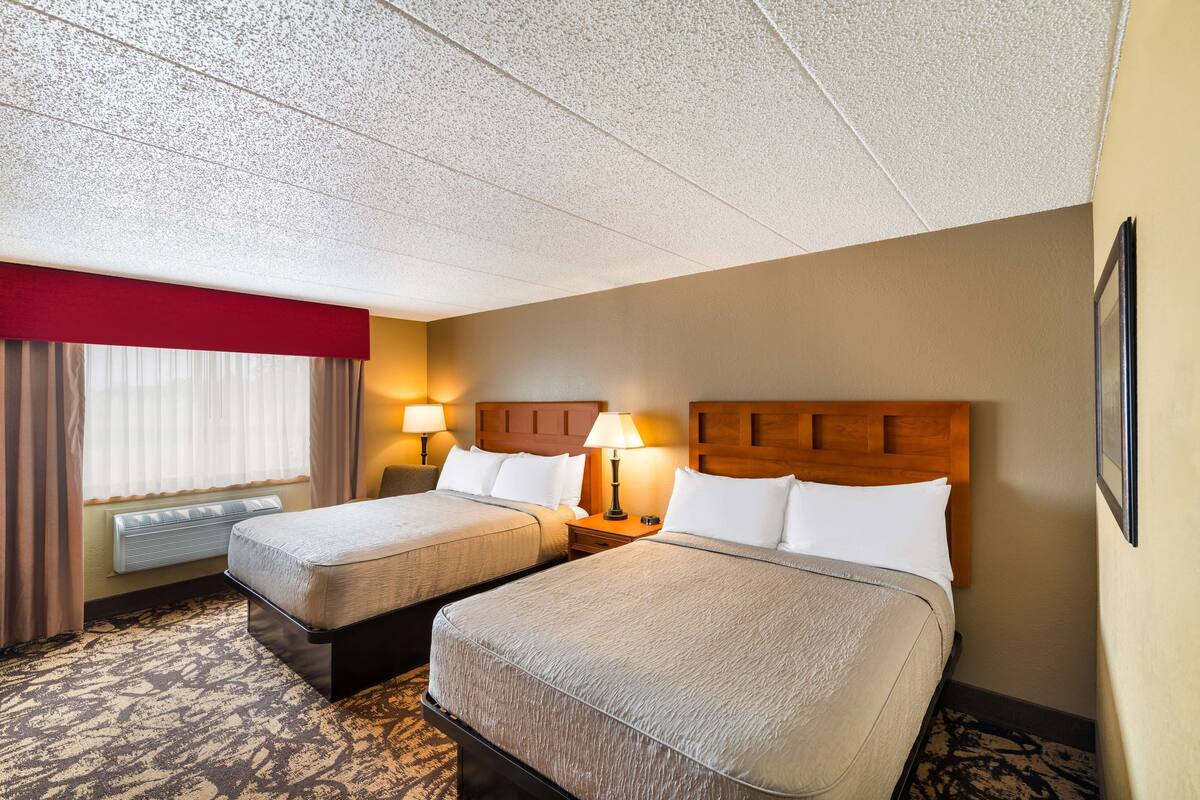 A hotel room with two beds, a red accent wall, a window with curtains, and a wall-mounted heater.