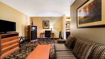 A hotel room with a sofa, a wooden table, a TV, a desk with a chair, a framed picture on the wall, and a carpeted floor.