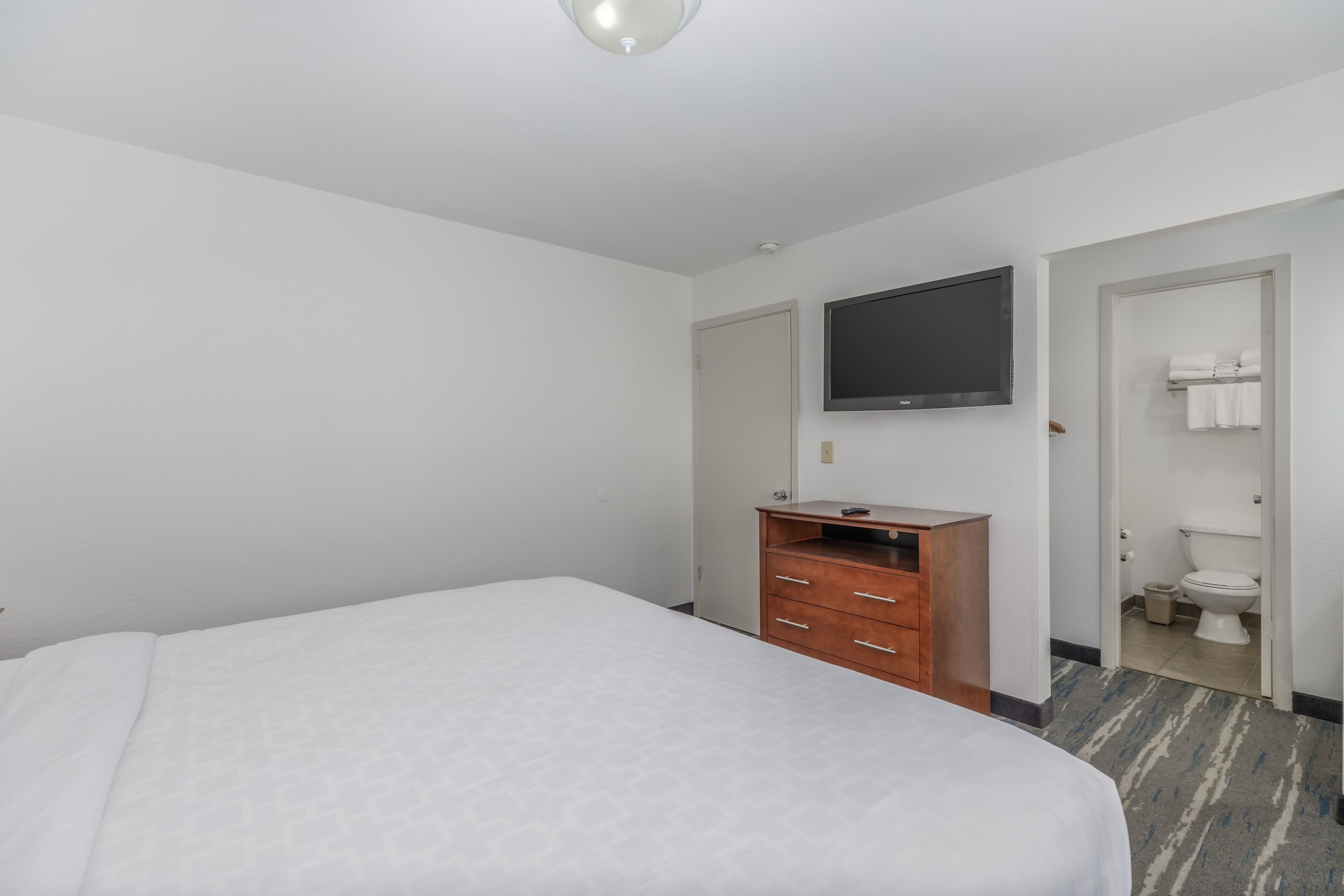 A hotel room with a large bed, a wooden dresser, a flat-screen TV mounted on the wall, and a bathroom visible through an open door.