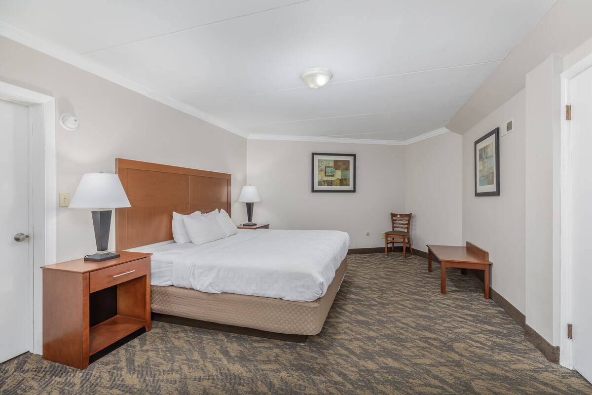 Ein Hotelzimmer mit einem Bett, zwei Nachttischen mit Lampen, einem Sessel und einem kleinen Tisch, sowie einem gerahmten Bild an der Wand.