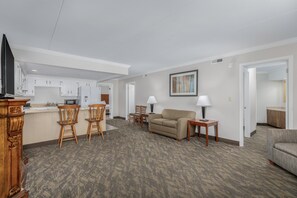 A modern living room with a sofa, two chairs, a coffee table, and a TV. There is a kitchen area with cabinets and a microwave.