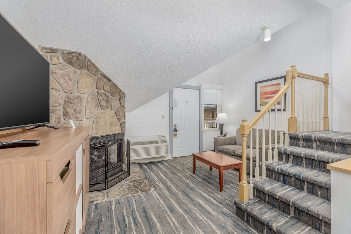 Ein Wohnzimmer mit einer Wand aus Stein, einem Kamin, einem Fernseher, einem Sofa, einem Couchtisch und einer Treppe mit Geländer.