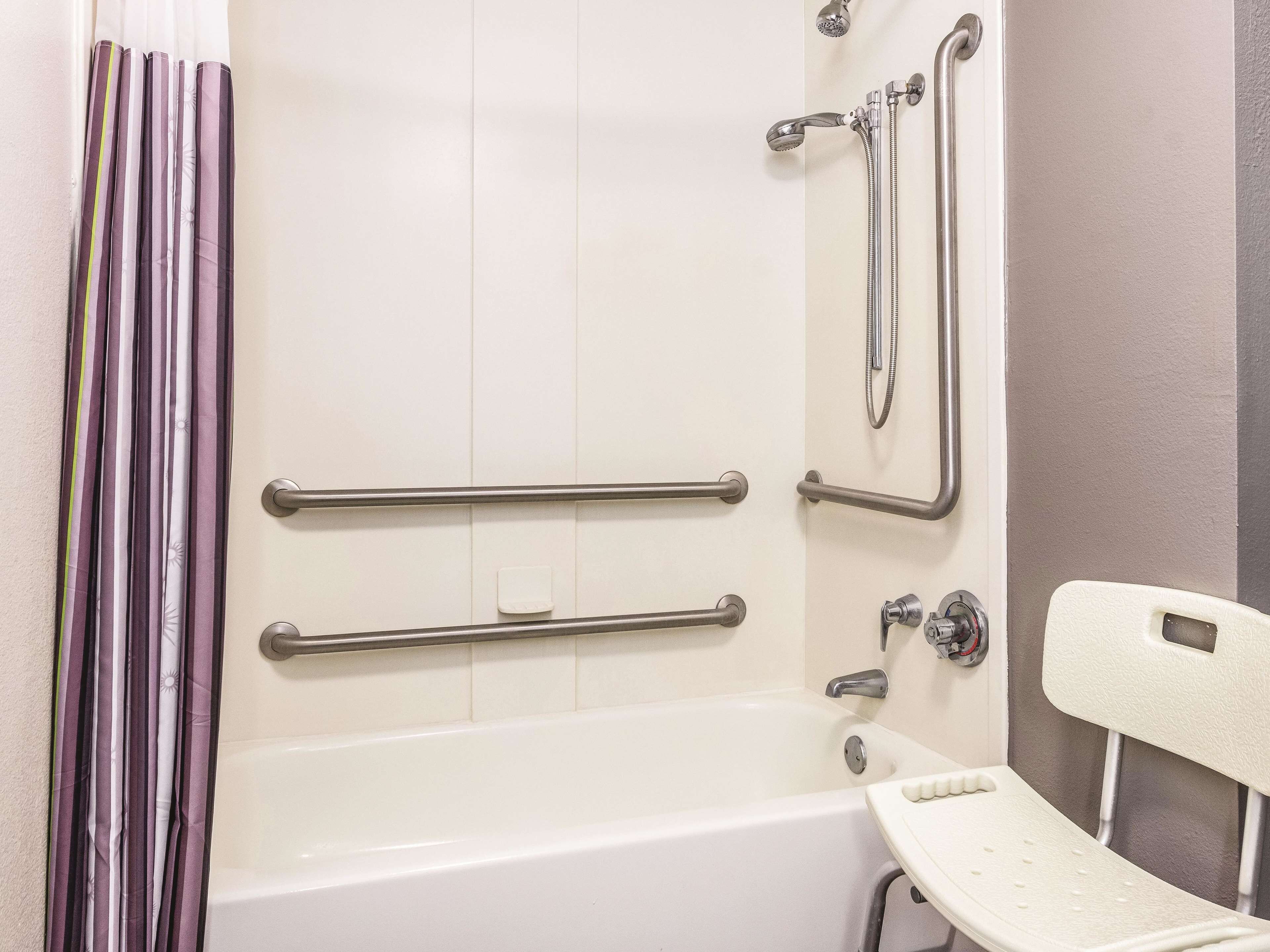 Un baño con bañera, barras de apoyo, ducha y una silla blanca.