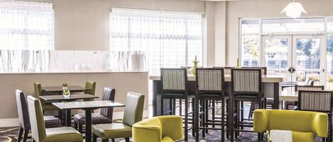 A hotel dining area with tables and chairs, large windows, and a view of the outdoors.
