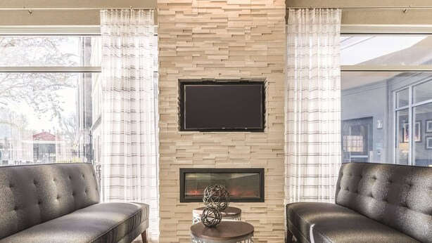 A modern living room with two leather sofas, a fireplace, a TV mounted on a stone wall, and large windows with curtains. - La Quinta Inn & Suites by Wyndham Alexandria Airport (Alexandria)