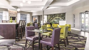 A modern hotel lobby with a central reception desk, multiple seating areas with purple and yellow chairs, a patterned carpet, and a large abstract painting on the wall. - La Quinta Inn & Suites by Wyndham Alexandria Airport (Alexandria)