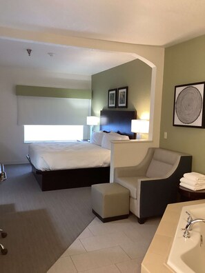 A hotel room with a double bed, a sofa bed, a small table, a chair, a desk, and a bathroom with a sink and mirror.