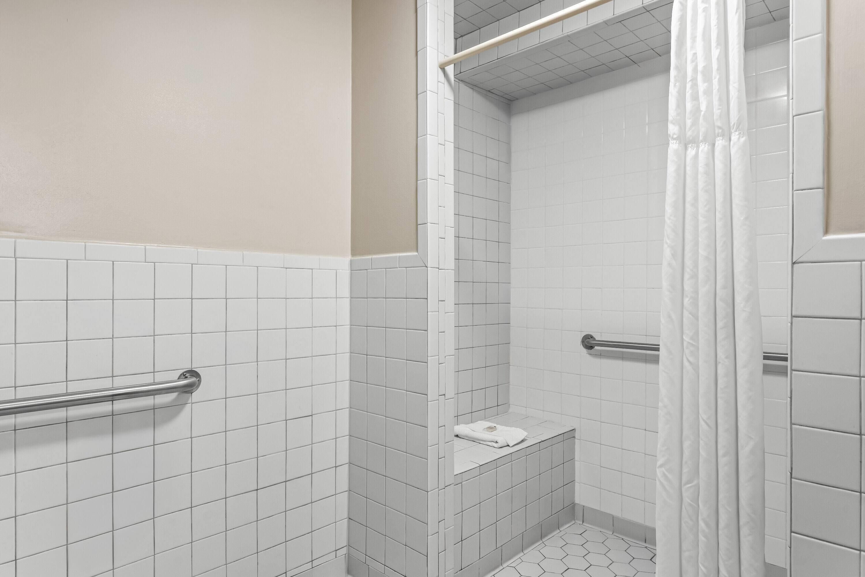 Un petit espace de douche carrelé, avec un rideau blanc, une serviette et une barre d’appui en métal.