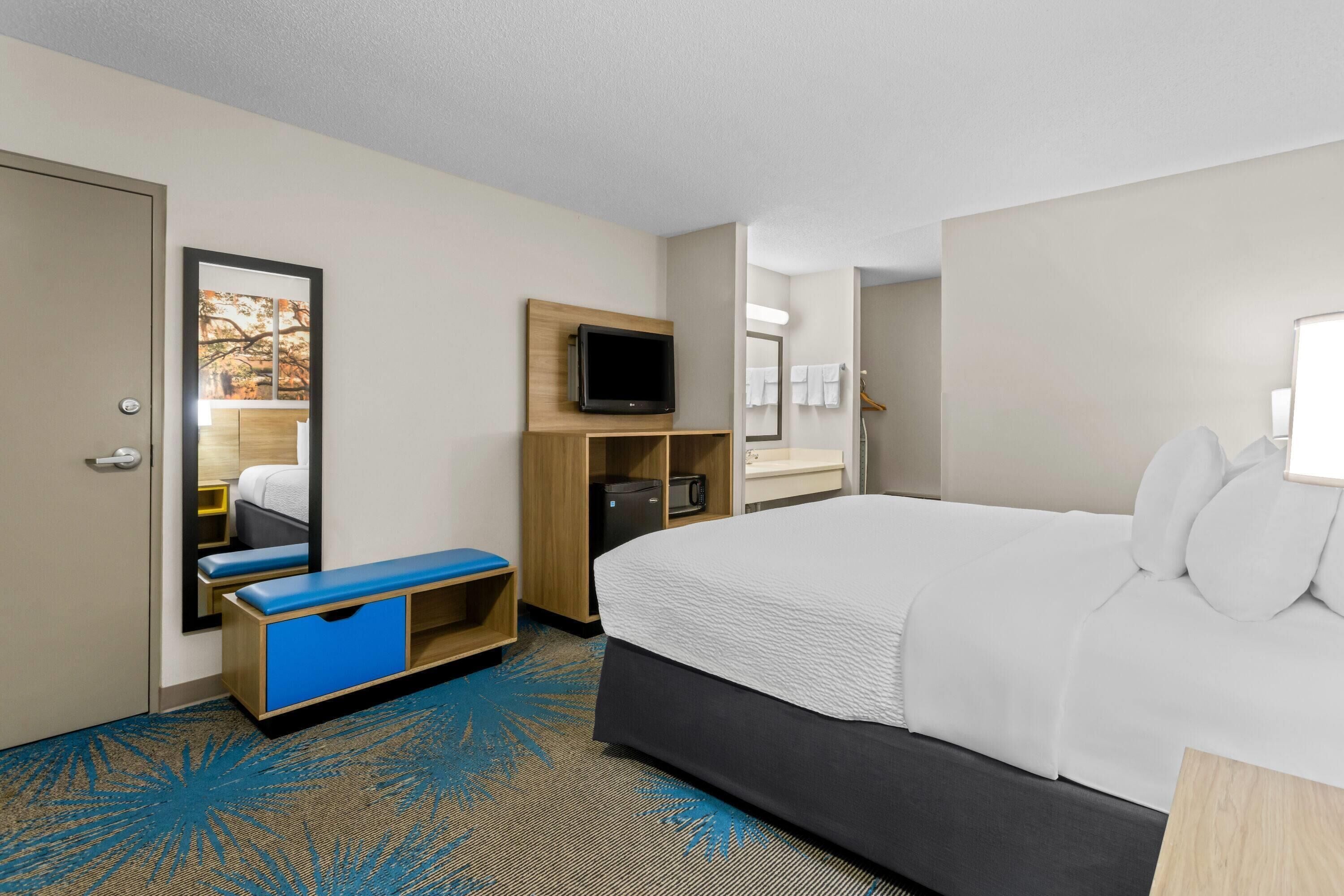 Una habitación de hotel con una cama, un escritorio con computadora, un televisor y un baño visible a través de una puerta abierta.