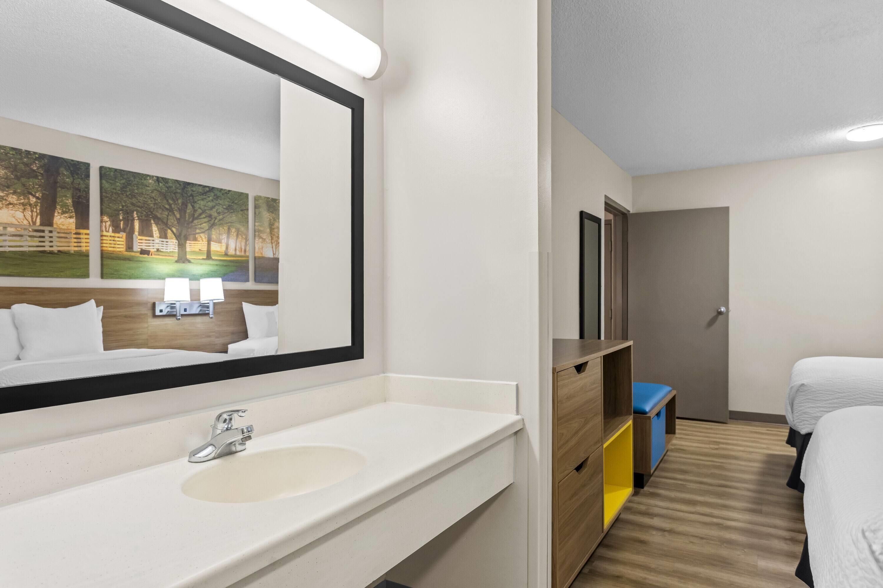Une chambre d’hôtel avec un grand miroir, un lavabo blanc, une armoire en bois, un tabouret bleu et deux lits.