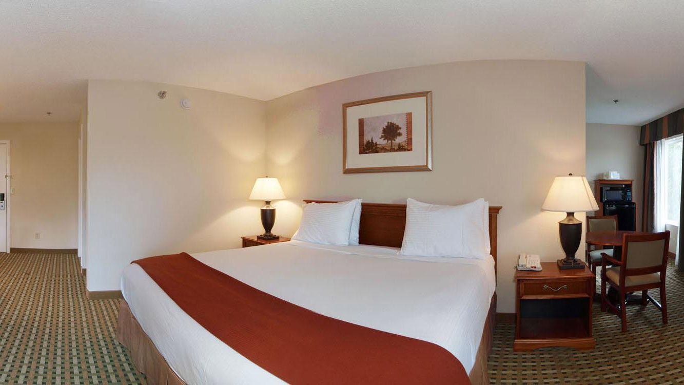 A hotel room with a large bed, two bedside tables with lamps, a chair, a desk with a chair, and a framed picture on the wall.