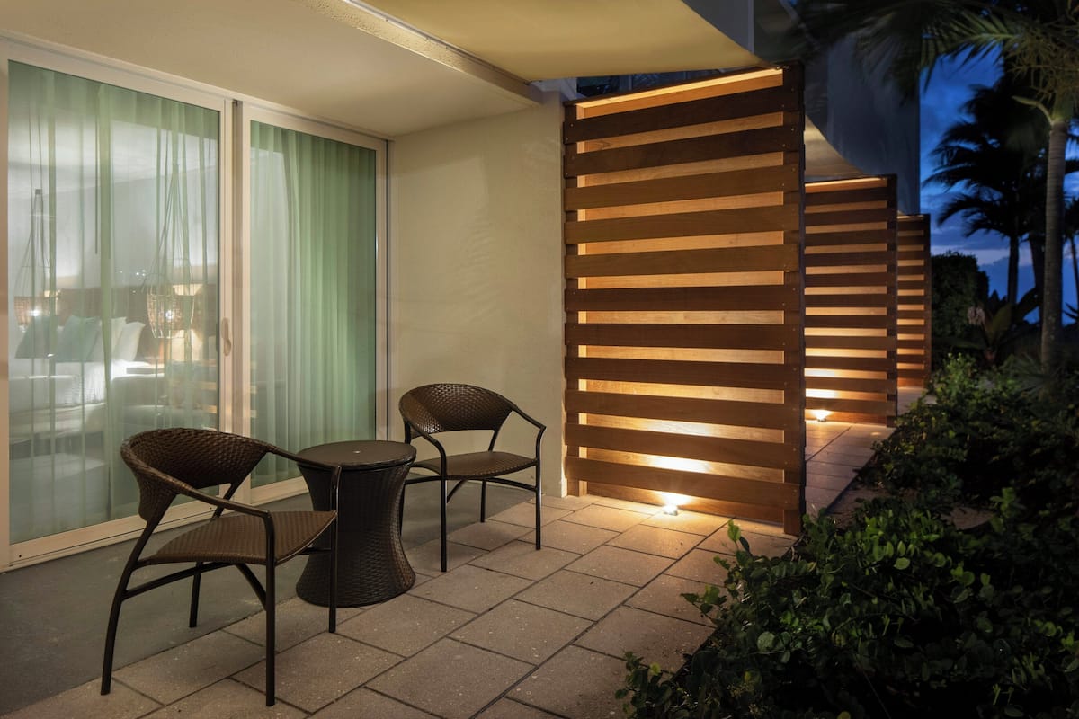 Une terrasse extérieure avec deux fauteuils en osier et une petite table ronde, un paravent en lattes de bois, et une vue sur un bâtiment doté de portes vitrées.
