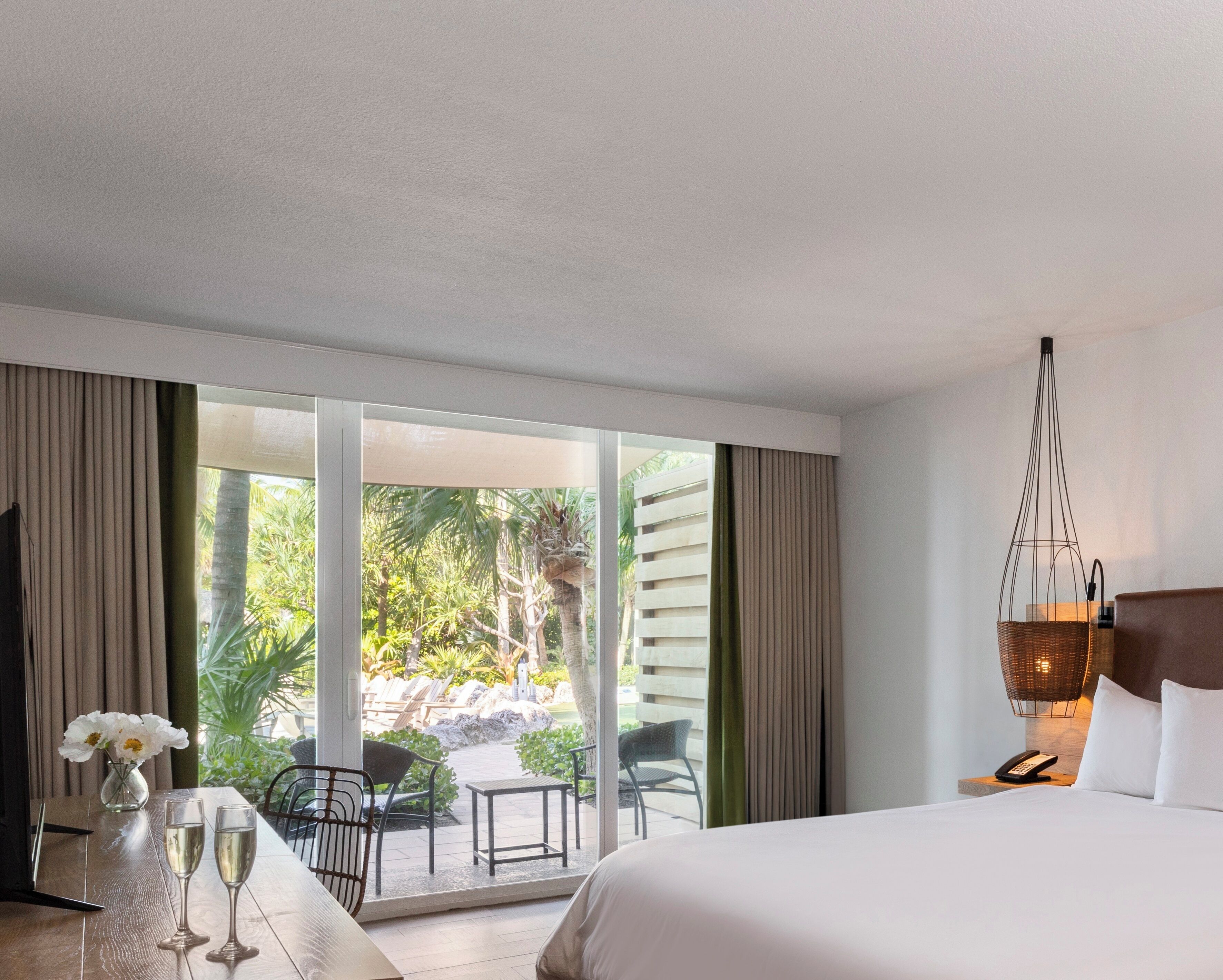 A hotel room with a bed, a table with two glasses of champagne, a vase with flowers, a chair, and a view of an outdoor area with palm trees and a pool.