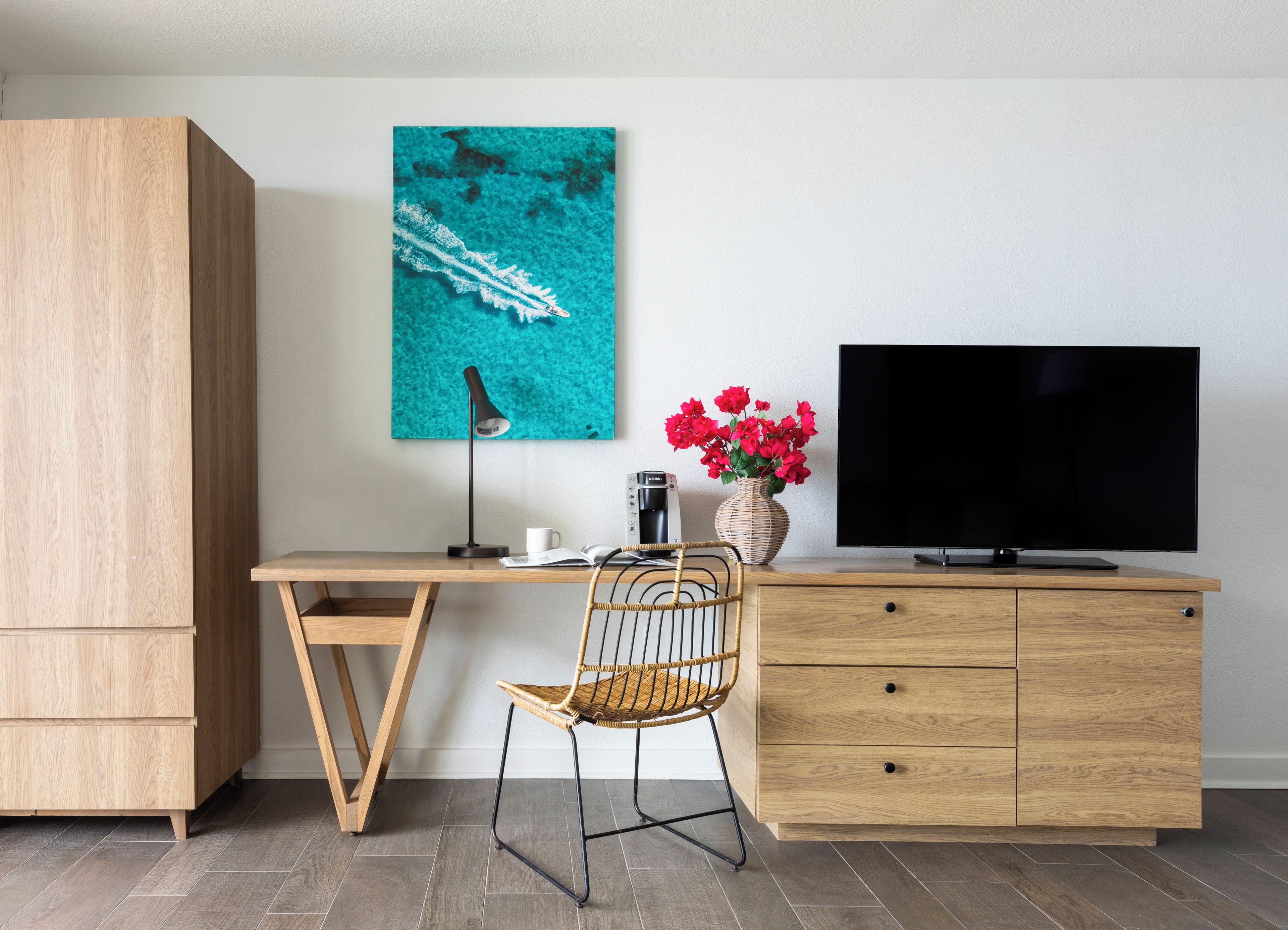 A modern office space with a wooden desk, a wicker chair, a television, a coffee maker, and a vase with flowers.