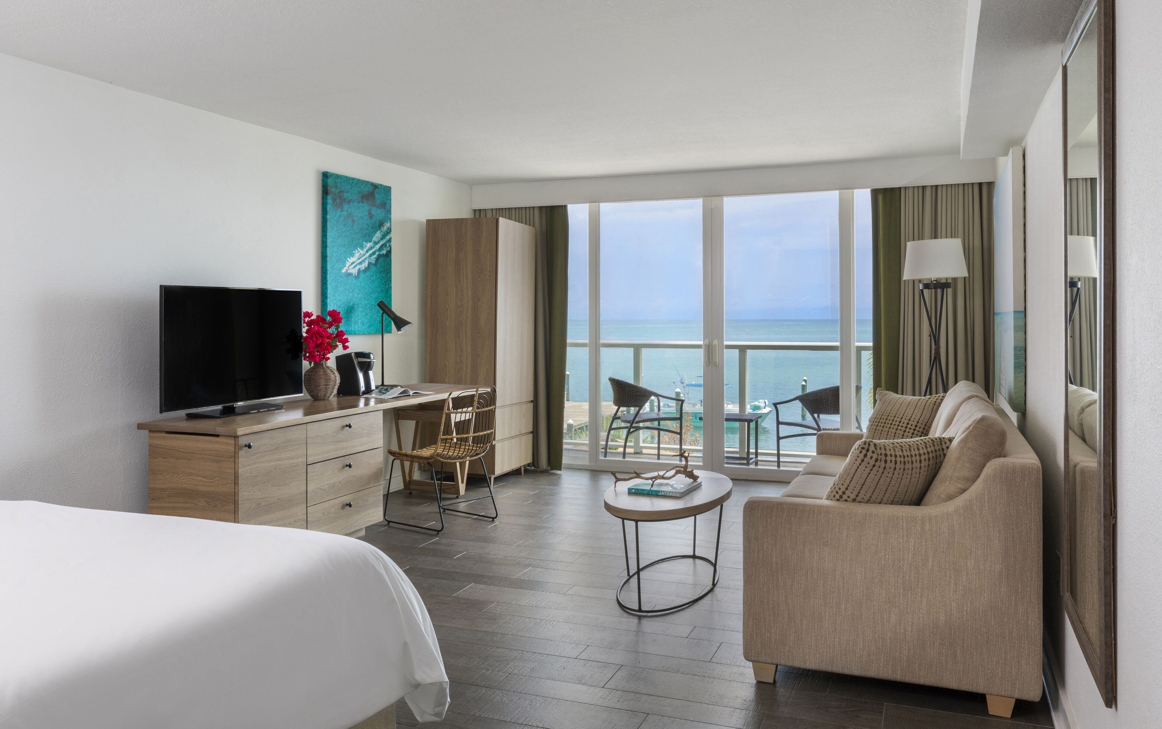 A hotel room with a bed, a desk with a TV, a chair, a sofa, a small table, and a view of the sea.