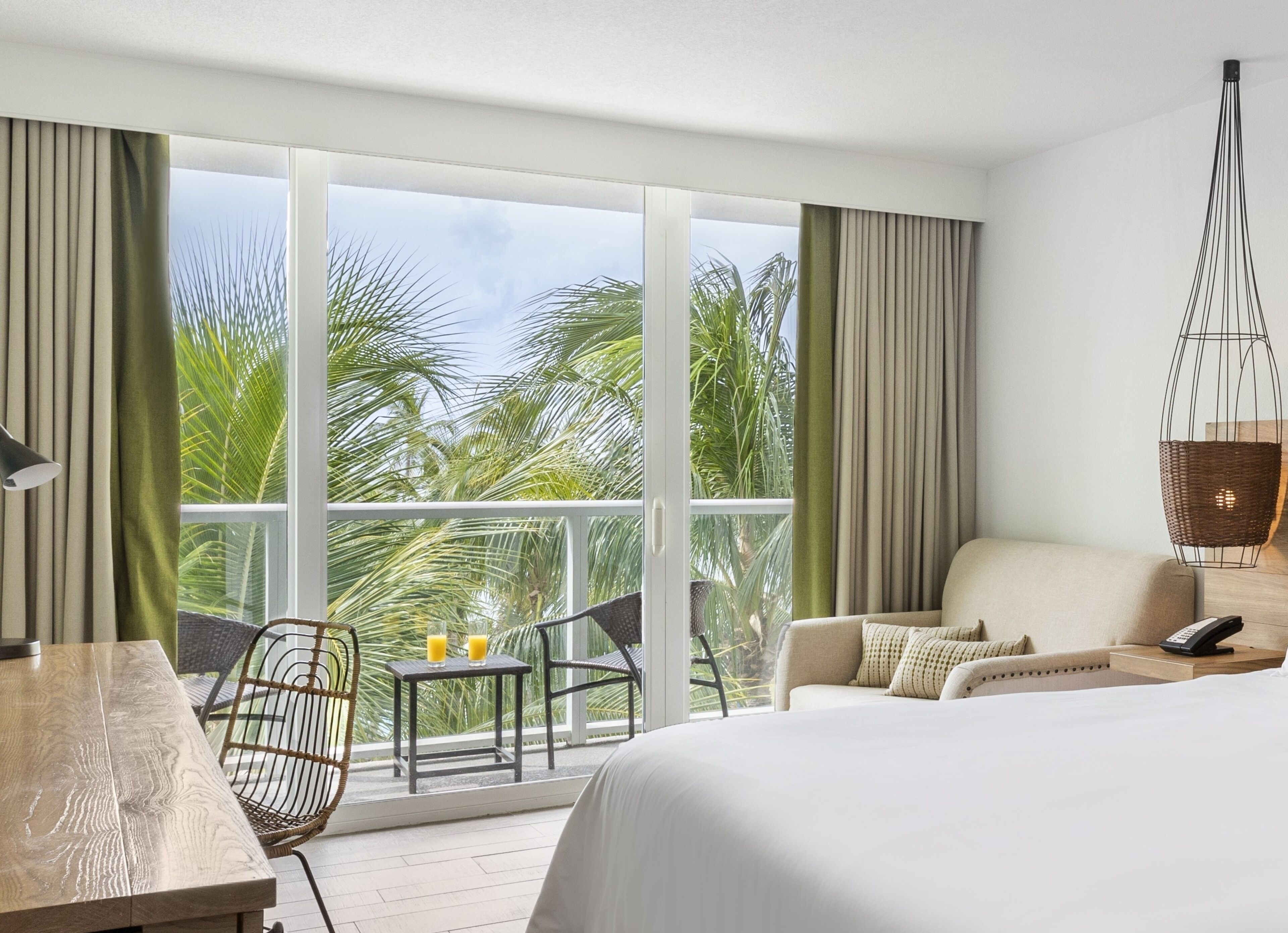 A hotel room with a large window, a bed, a table, a chair, and a hanging light fixture.