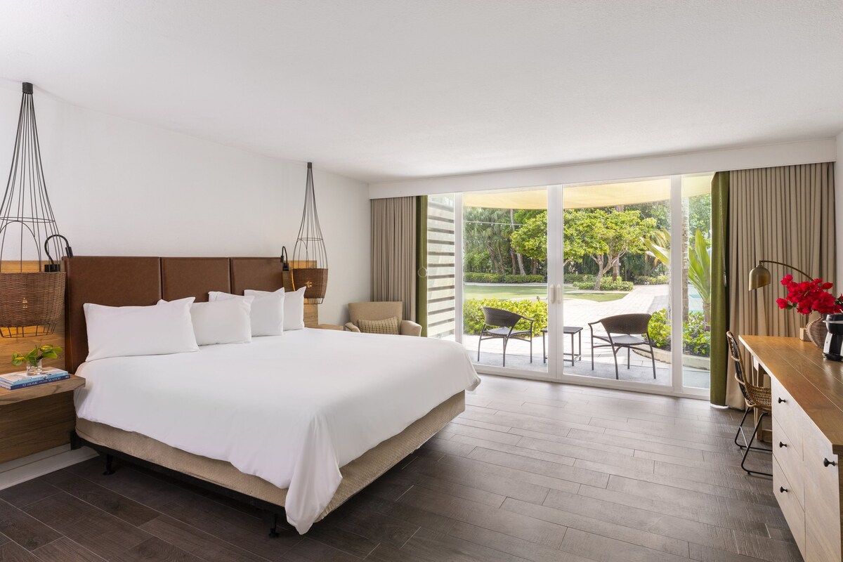 A hotel room with a large bed, a wooden headboard, a nightstand with a lamp, a desk with a chair, and a view of the garden through the sliding glass door.