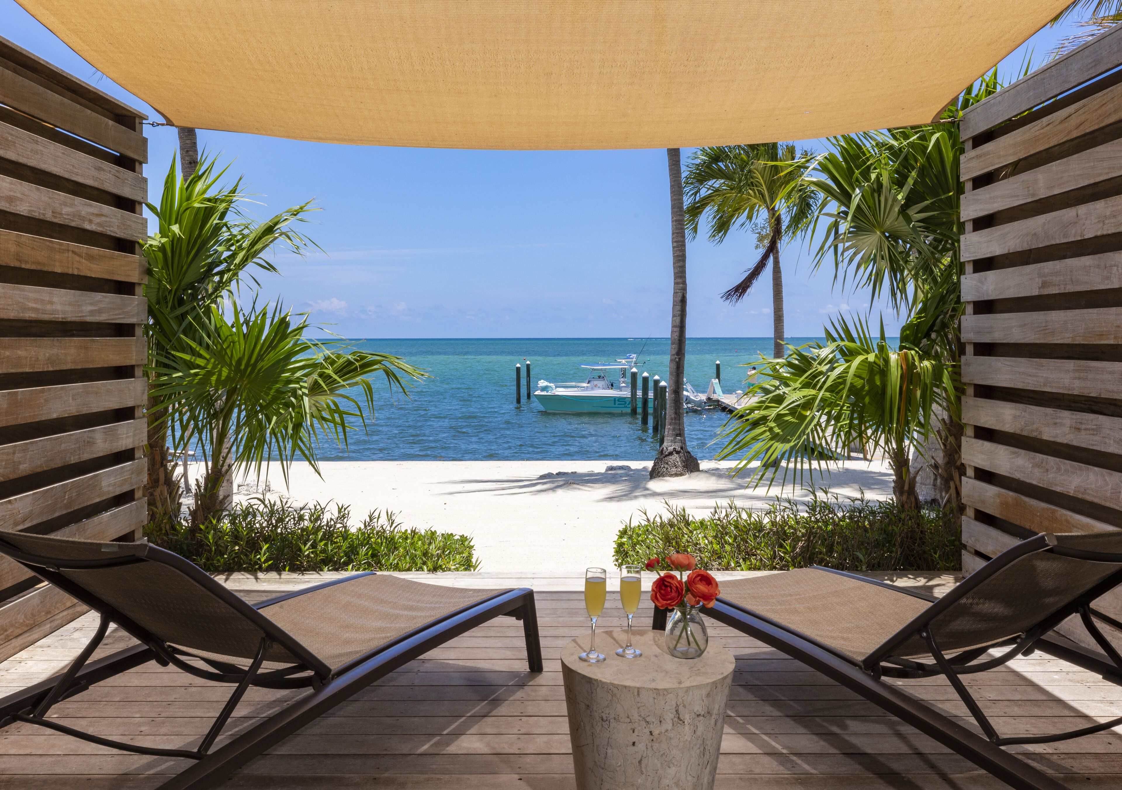 A wooden deck with two lounge chairs, a table with drinks and flowers, and a view of the beach and ocean.