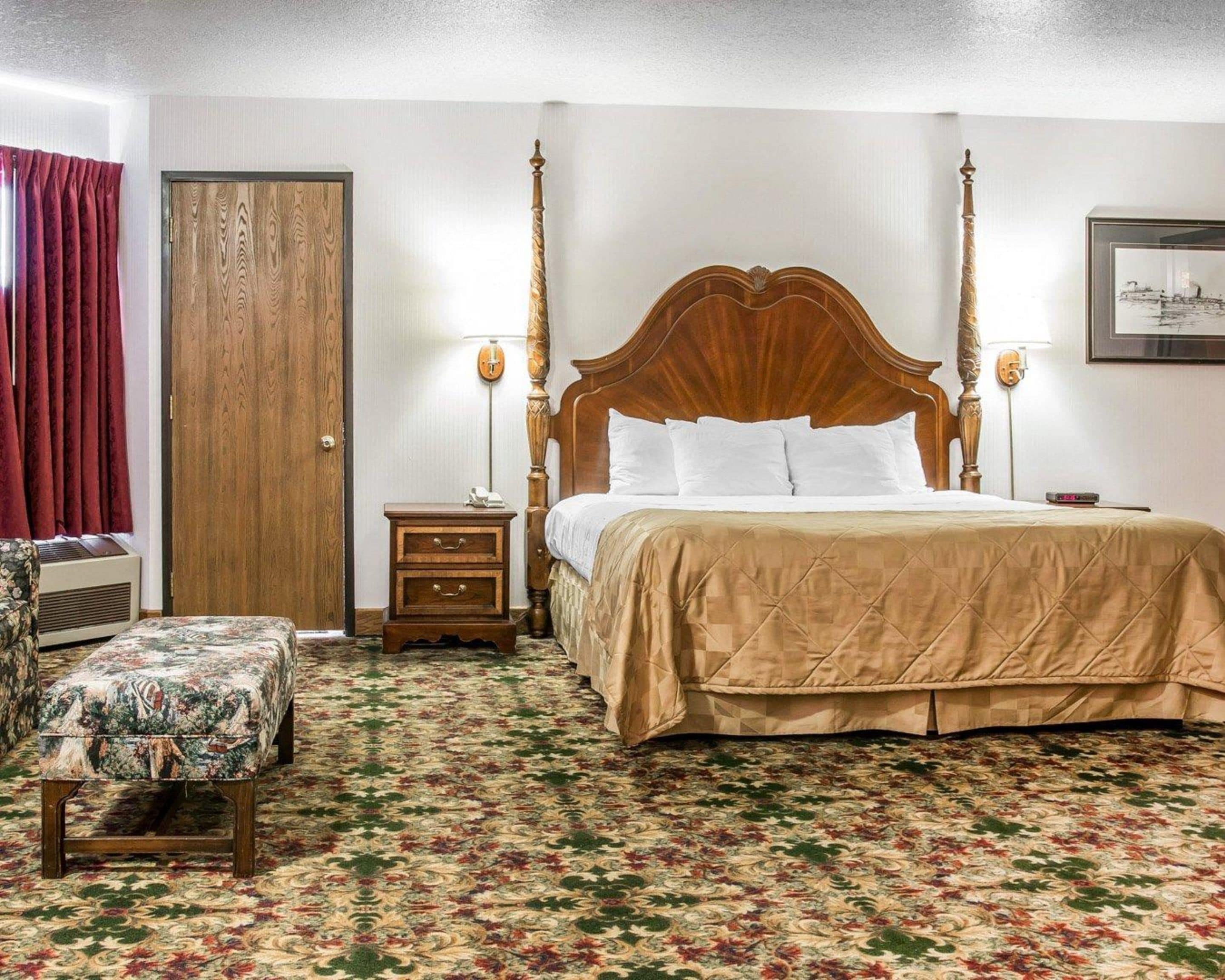 Una cama con dosel y tablero de madera, una mesita de noche con lámpara, un banco estampado con motivos florales y una alfombra con diseño.