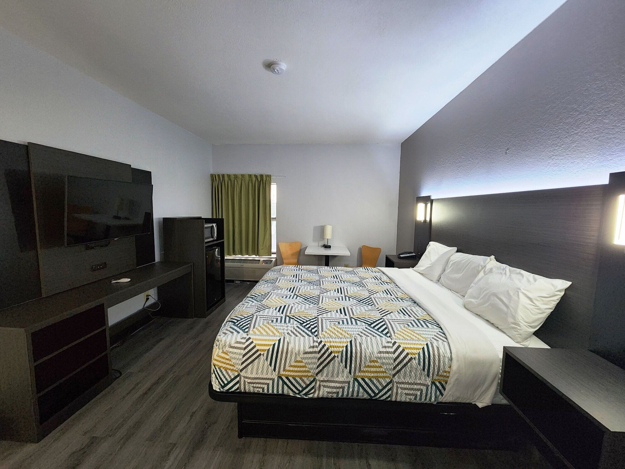 A hotel room with a large bed, a flat-screen TV, a desk with a lamp, a chair, and a small table.