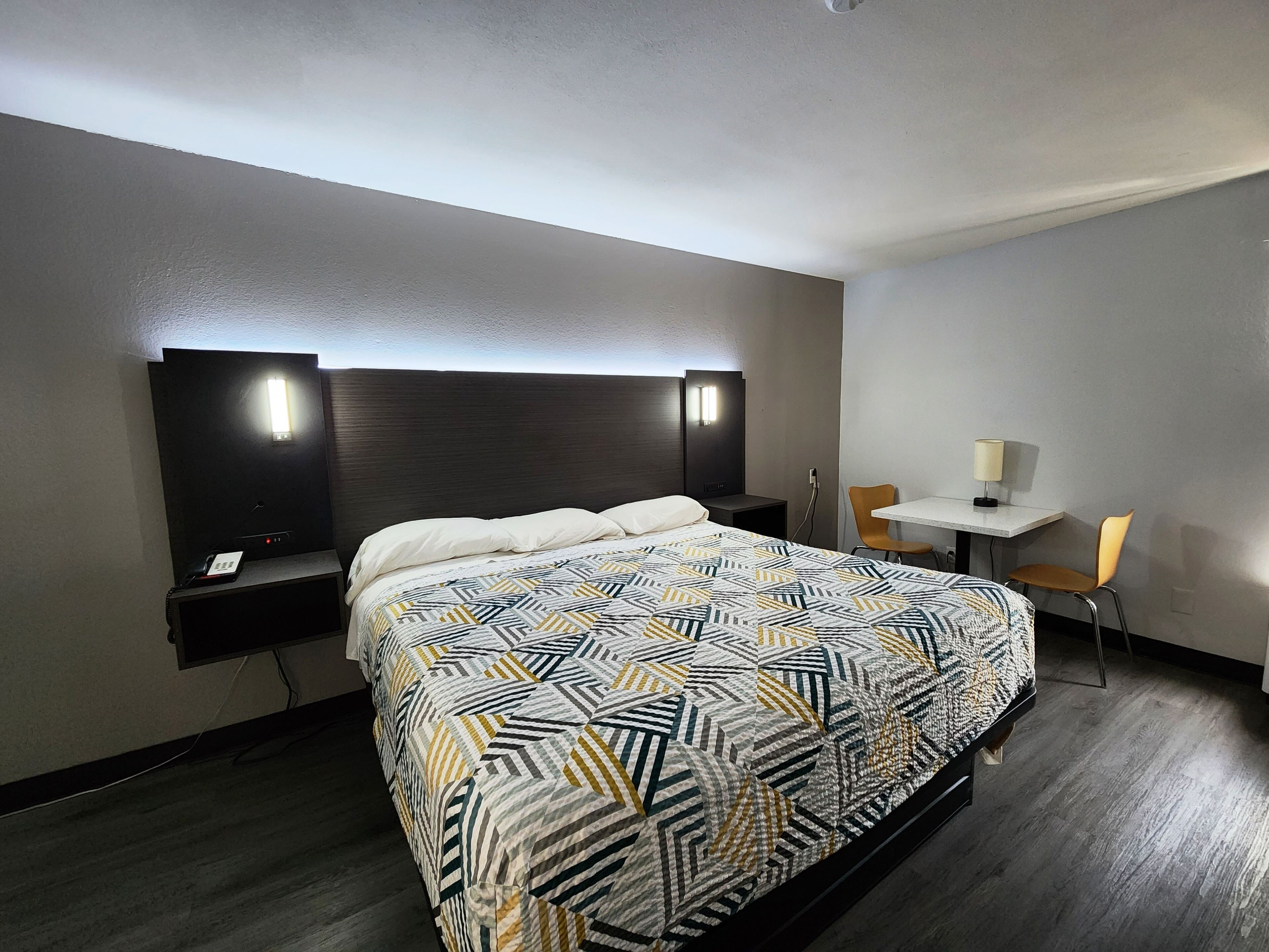 A hotel room with a large bed, a desk with a chair, and a small table with a lamp.