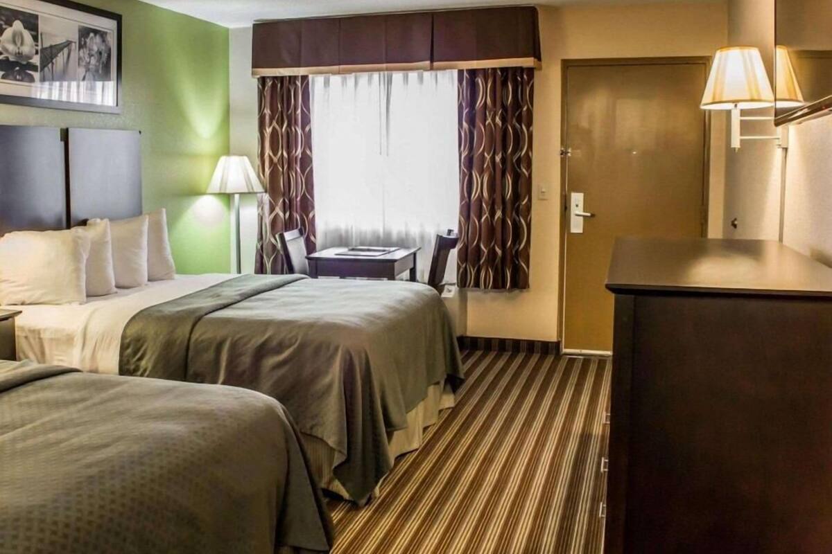 Ein Hotelzimmer mit zwei Betten, einem Schreibtisch, einem Stuhl, einer Lampe, einer Tür und einem Fenster mit Vorhängen.