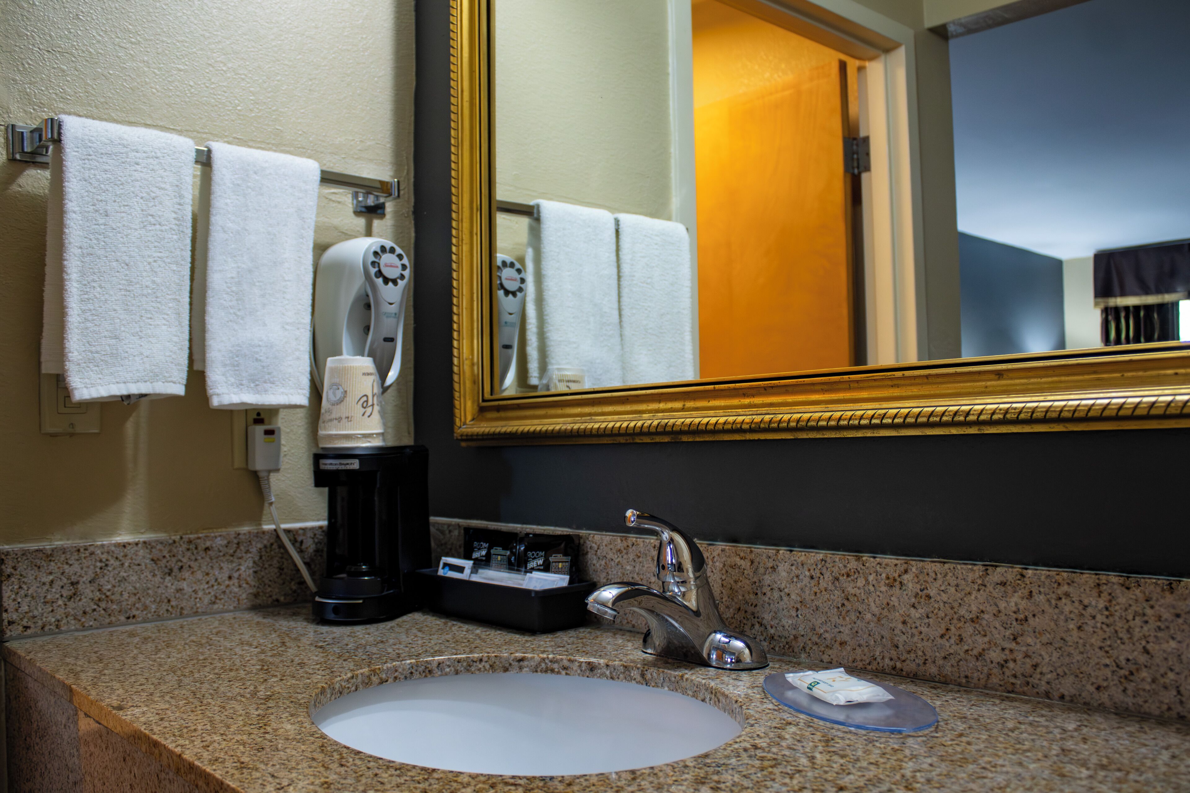 A hotel bathroom with a granite countertop, a sink, a mirror, and a coffee maker.