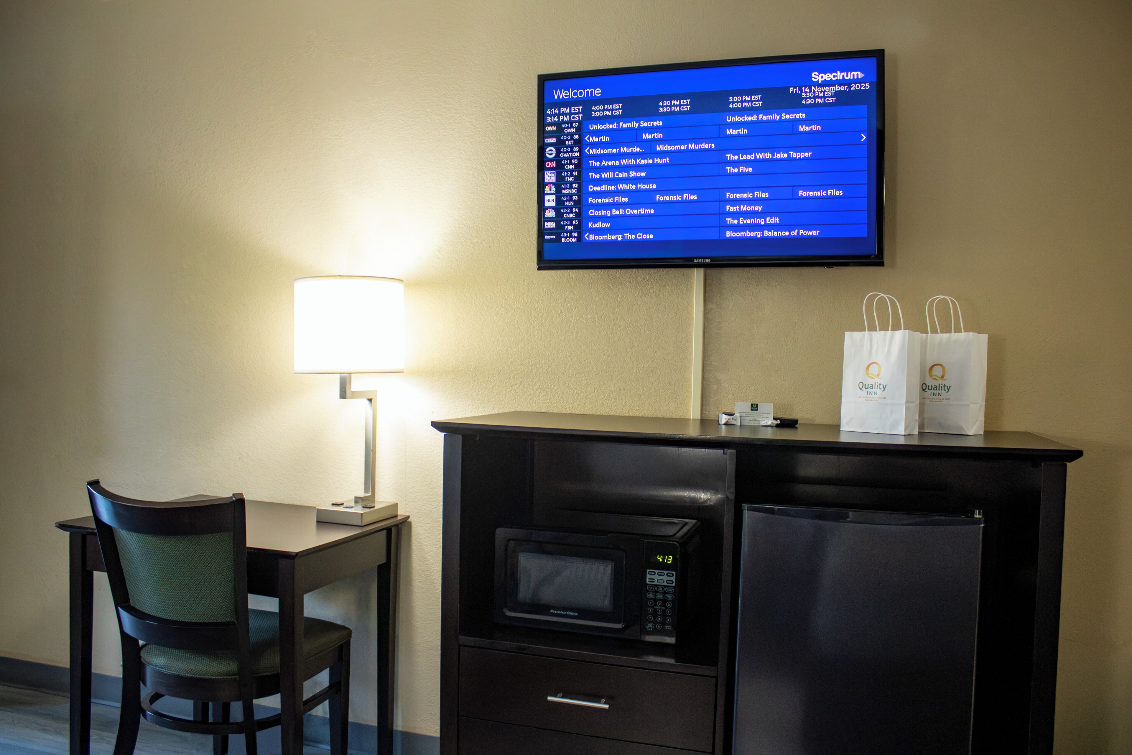 Habitación de hotel con televisión de pantalla plana mostrando una pantalla de bienvenida, un escritorio con lámpara, un microondas y un refrigerador. Hay dos bolsas de compras sobre el refrigerador.