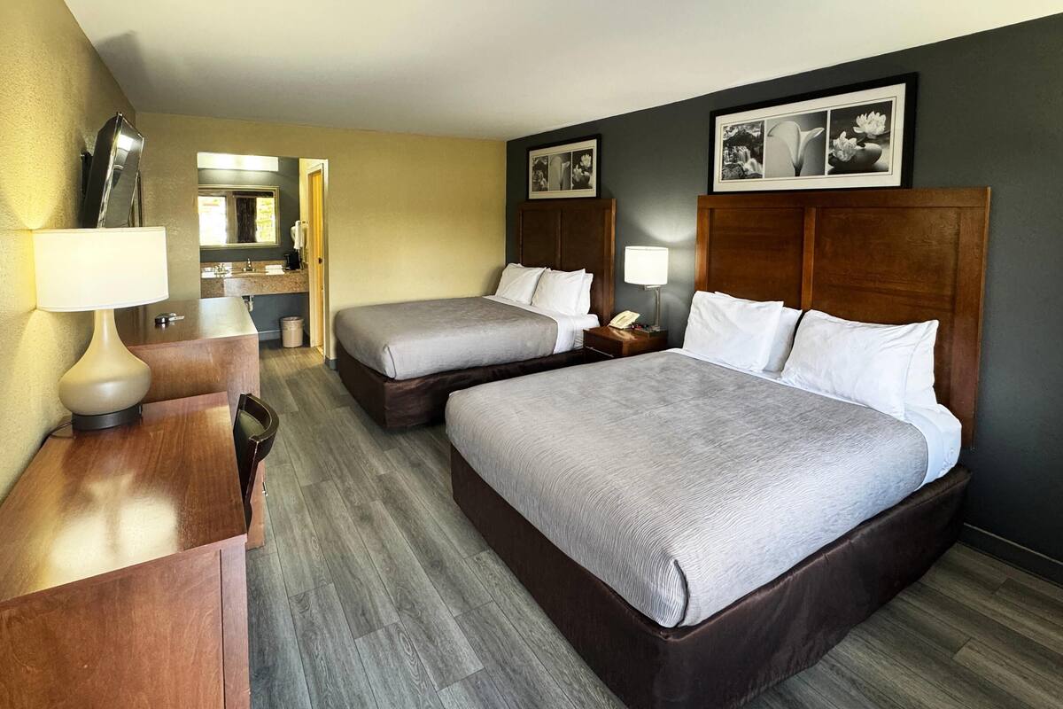 Ein Hotelzimmer mit zwei Betten, einem Schreibtisch, einer Lampe, einem Spiegel und einem durchgegangene Tür erkennbaren Badezimmer.