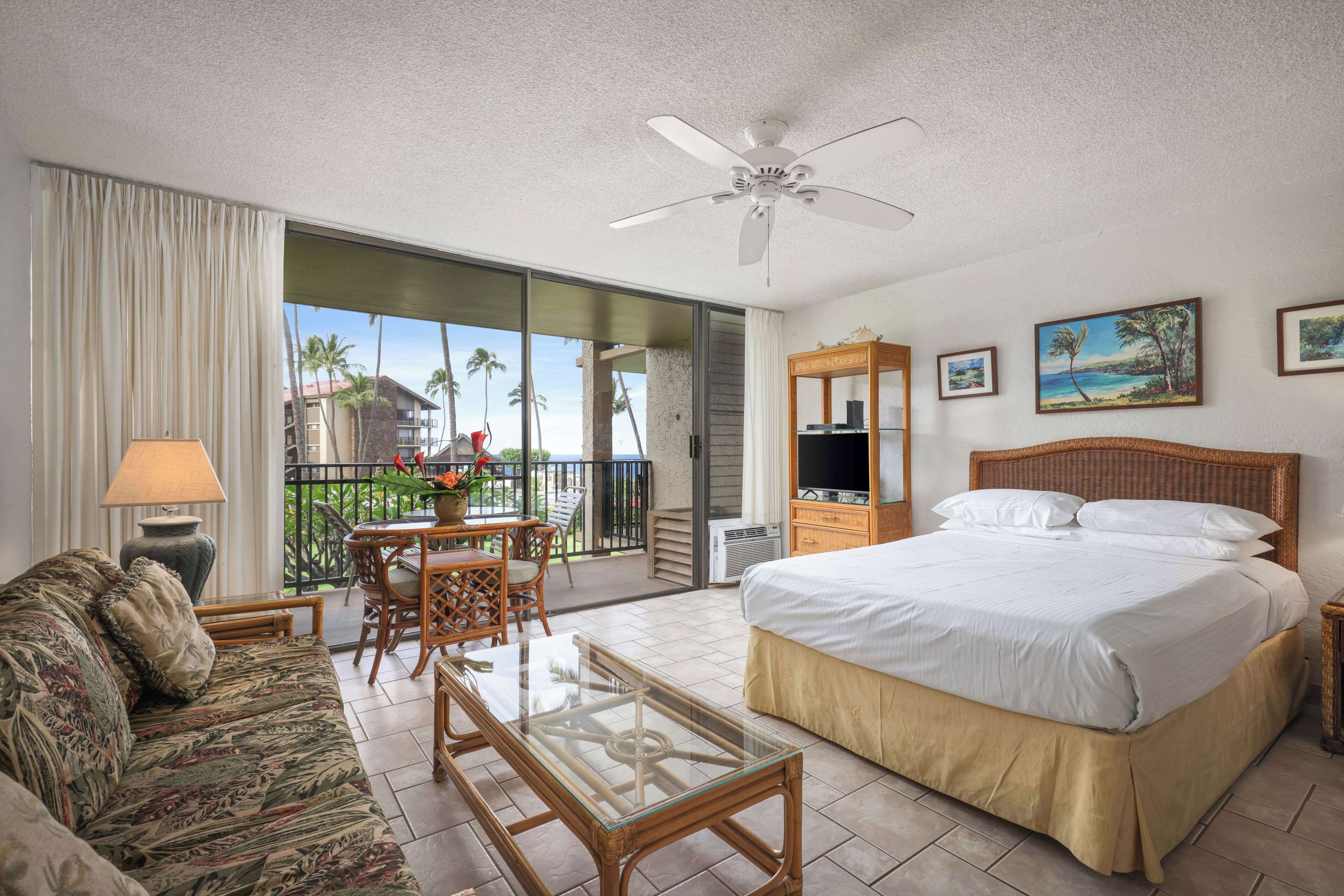 A hotel room with a bed, a sofa, a glass coffee table, a ceiling fan, a TV, and a balcony with a view of palm trees and buildings.