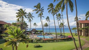 A resort with a pool area surrounded by palm trees, a view of the ocean, and a well-maintained lawn.