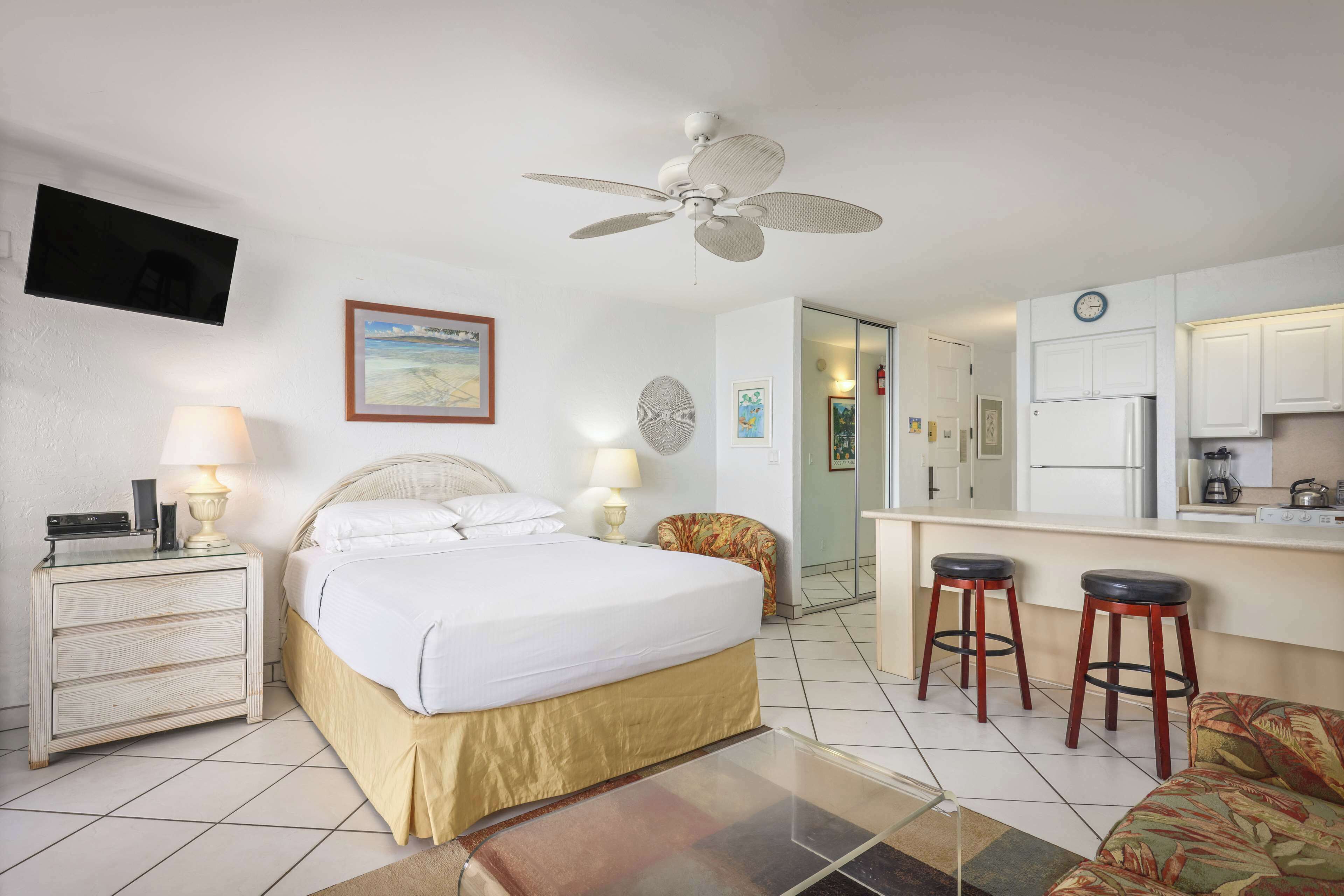 A hotel room with a bed, bedside lamps, a ceiling fan, a TV, a kitchen area with stools, and a door leading to another room.
