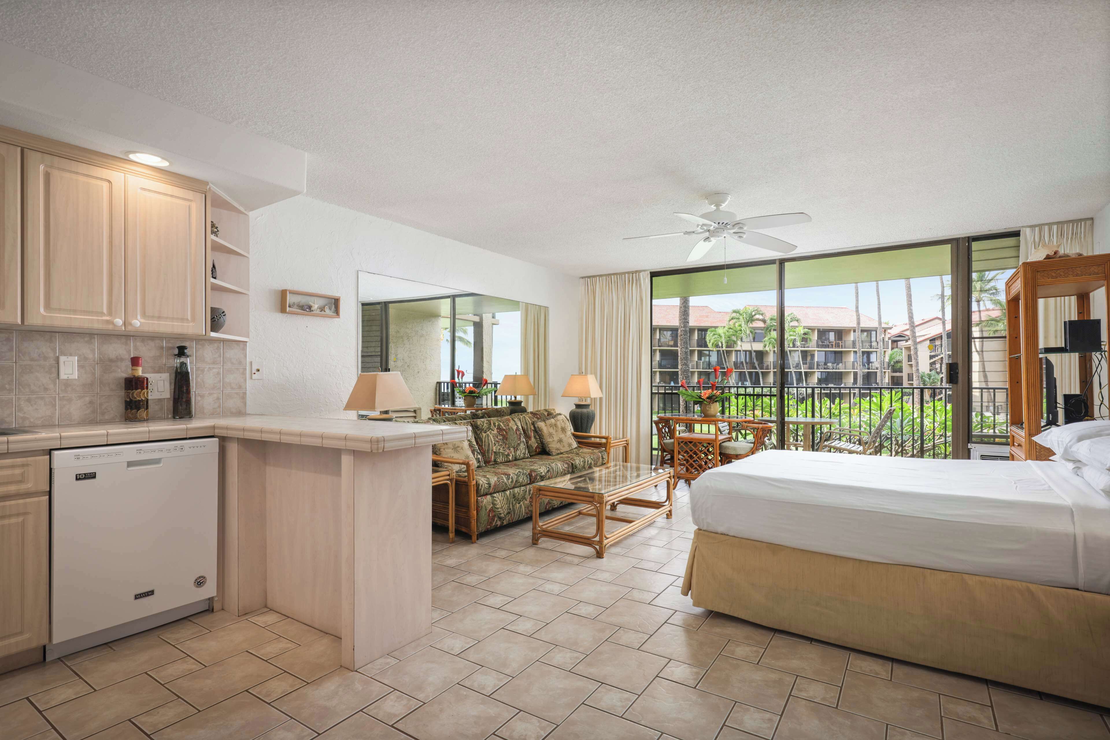 A hotel room with a kitchenette, a bed, a sofa, a dining area, and a balcony with a view of palm trees.
