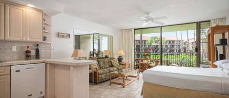 A hotel room with a kitchenette, a bed, a sofa, a dining area, and a balcony with a view of palm trees.