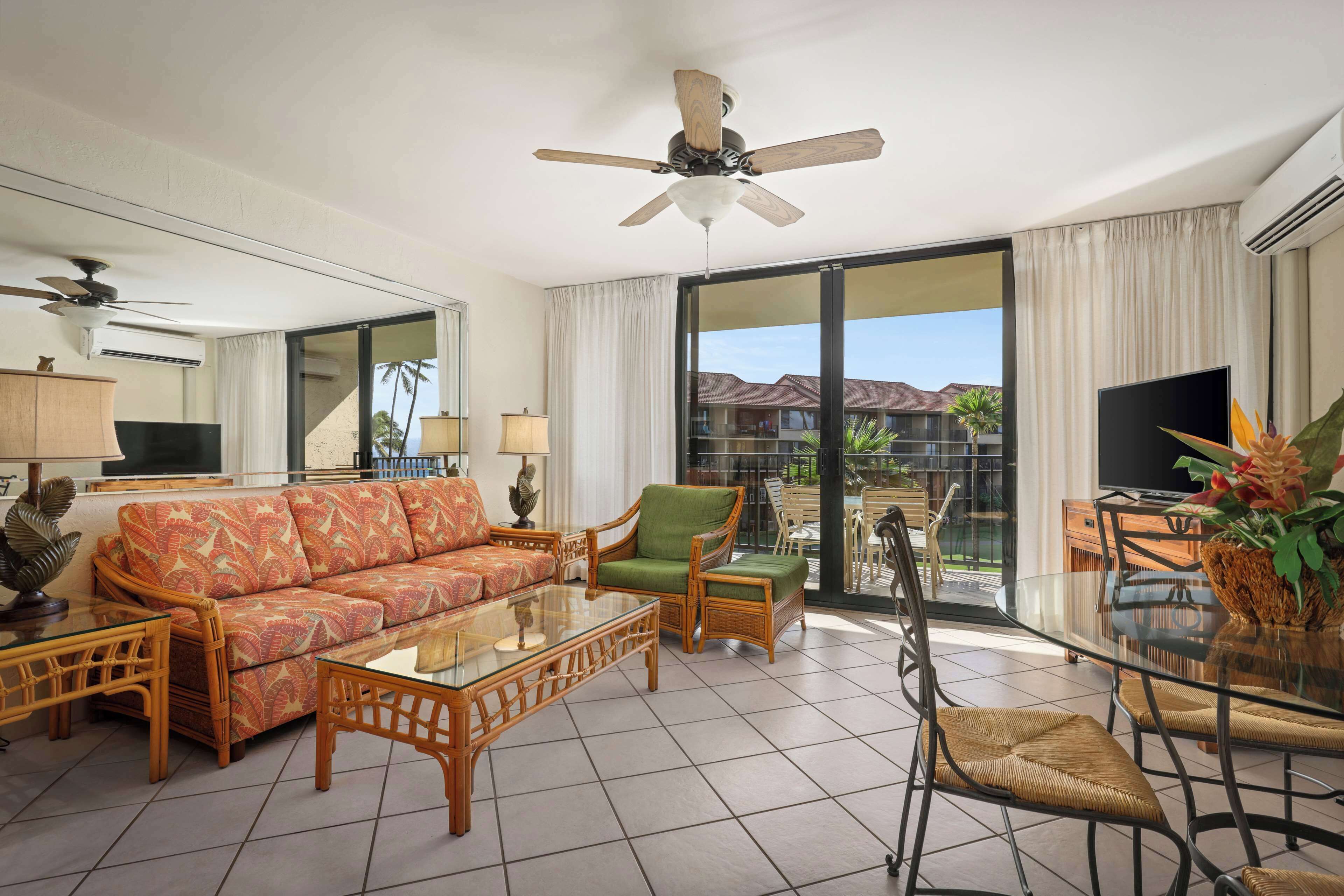 A spacious living room with a sofa, chairs, a glass coffee table, a ceiling fan, and a large window offering a view of palm trees and a building.